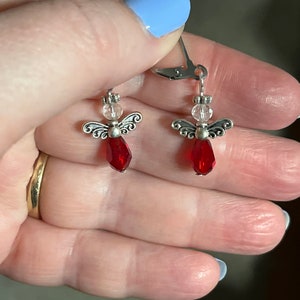 May include: A pair of silver-toned angel earrings. Each earring features a red, teardrop-shaped bead as the angel's body, a clear bead for the head, and silver wings. The earrings are held by a hand with light blue nail polish.