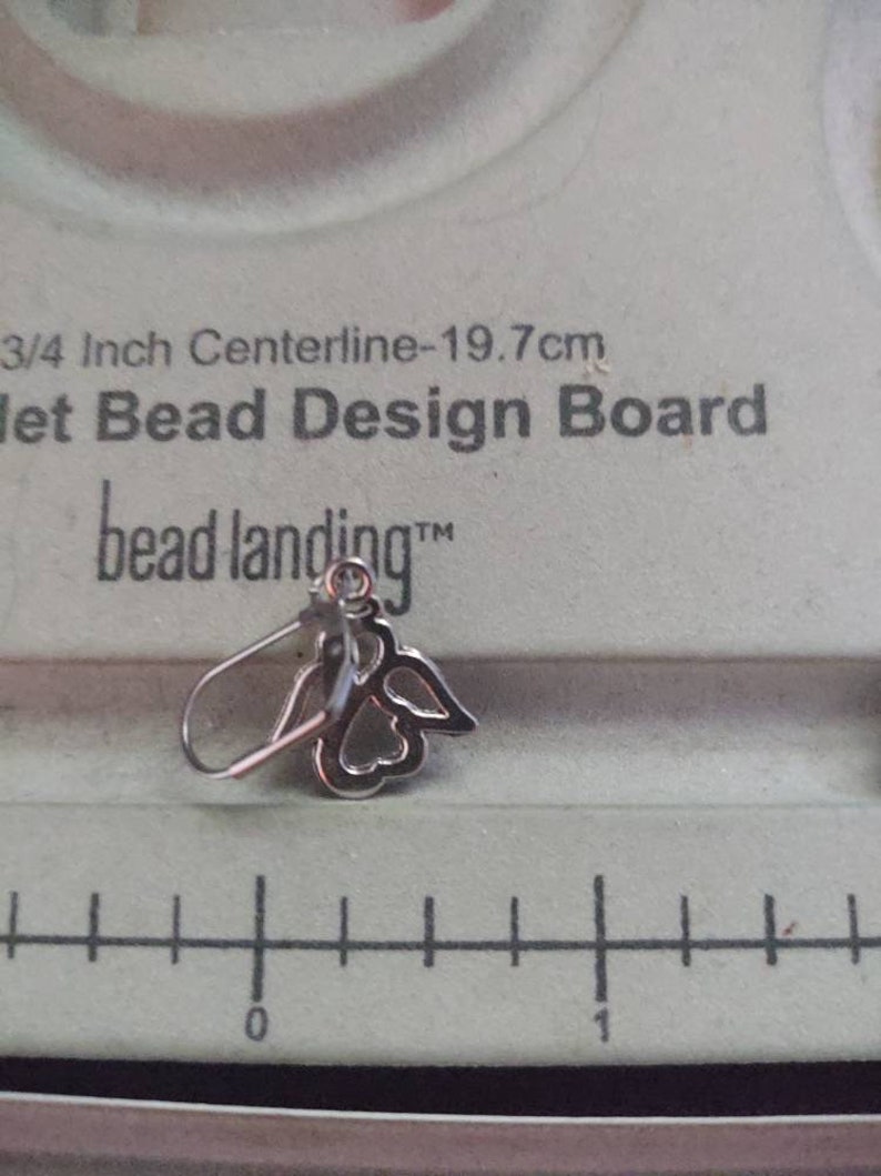 May include: A silver angel wing earring with a clear crystal in the center. The earring is on a white bead design board with the text "3/4 Inch Centerline-19.7cm Jet Bead Design Board bead landing&trade;".