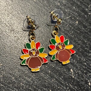 May include: A pair of gold-tone dangle earrings featuring a cartoon turkey design. The turkey is brown with red, yellow, and green feathers. The earrings have clear plastic backs.