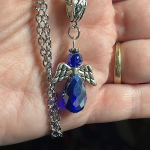 Pu&ograve; includere: Una collana a catena d'argento con un ciondolo a forma di angelo in cristallo blu. L'angelo ha ali d'argento e un corpo in cristallo blu sfaccettato.