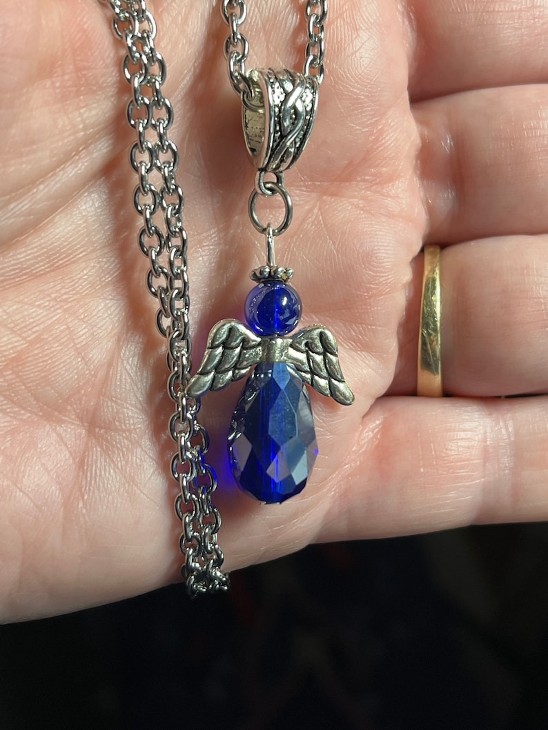 Pu&ograve; includere: Una collana a catena d'argento con un ciondolo a forma di angelo in cristallo blu. L'angelo ha ali d'argento e un corpo in cristallo blu sfaccettato.