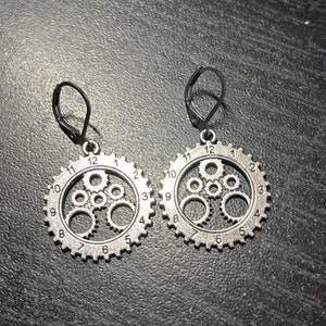 May include: A pair of silver earrings with a gear design. Each earring features a circular gear with smaller gears inside, and numbers around the outer edge.