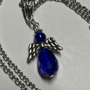 Pu&ograve; includere: Una collana a catena d'argento con un ciondolo a forma di angelo in vetro blu. L'angelo ha ali d'argento e un corpo a forma di lacrima.