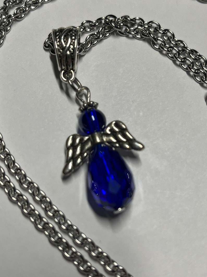 Pu&ograve; includere: Una collana a catena d'argento con un ciondolo a forma di angelo in vetro blu. L'angelo ha ali d'argento e un corpo a forma di lacrima.