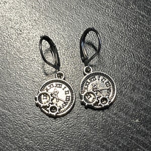 May include: A pair of silver-toned steampunk-style earrings. Each earring features a circular clock face design with Roman numerals and gear details. The earrings have black lever-back closures.