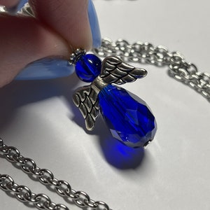 Pu&ograve; includere: Una collana a catena d'argento con un ciondolo a forma di angelo in cristallo blu. L'angelo ha ali d'argento e un corpo in cristallo blu.