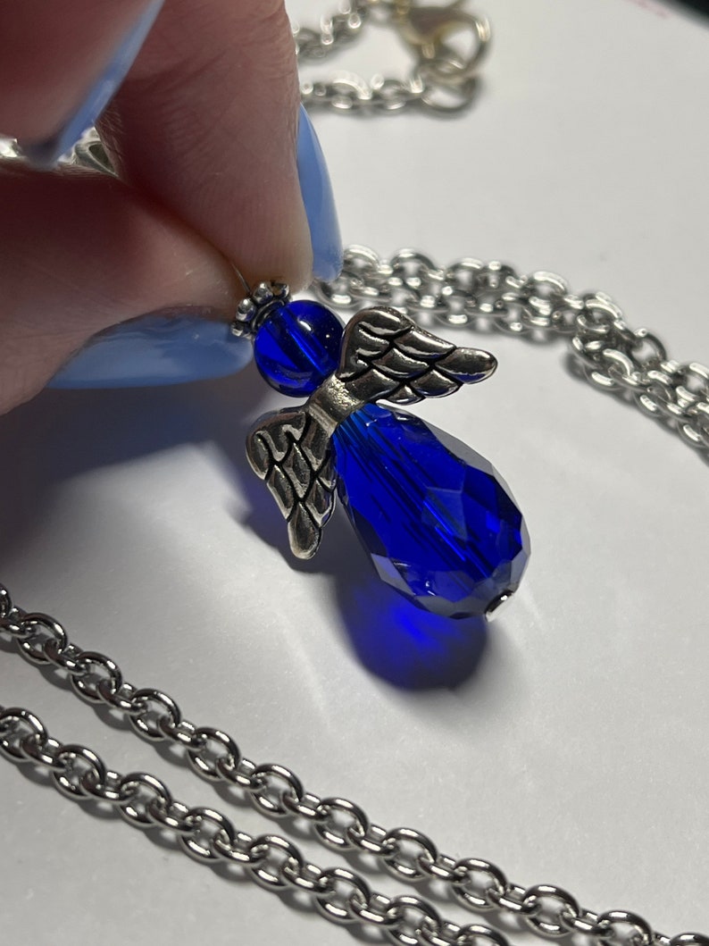 Pu&ograve; includere: Una collana a catena d'argento con un ciondolo a forma di angelo in cristallo blu. L'angelo ha ali d'argento e un corpo in cristallo blu.