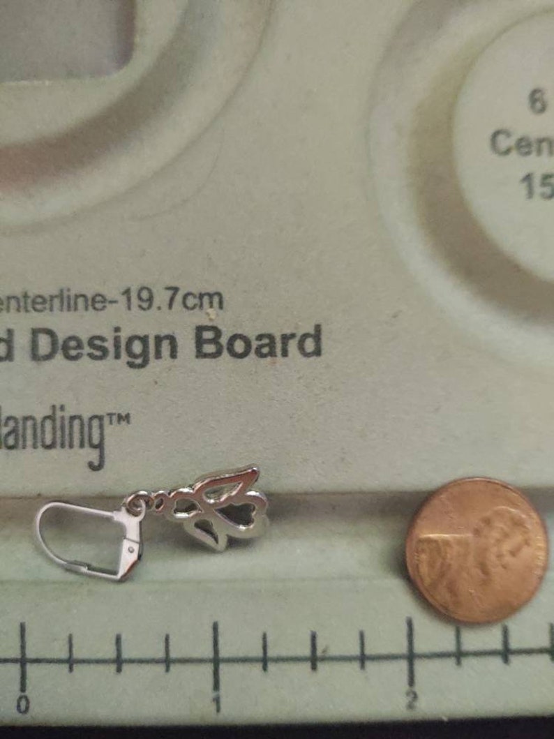 May include: Silver angel wing earring with a hook closure. The earring is about 1 inch long and is shown next to a US penny for size comparison.