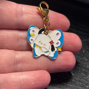 May include: A gold-colored charm with a white butterfly design. The butterfly has blue wings with purple and yellow accents. The charm has a gold clasp.