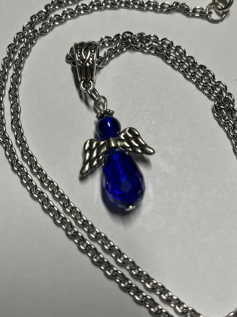 Pu&ograve; includere: Una collana a catena d'argento con un ciondolo a forma di angelo in cristallo blu. L'angelo ha ali d'argento e un corpo a forma di lacrima.