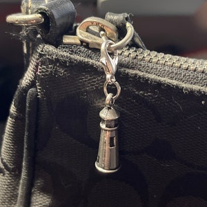 May include: A silver charm in the shape of a lighthouse hangs from a black zipper pull on a black bag.