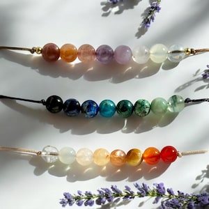 Könnte beinhalten: Drei Perlenketten mit unterschiedlich farbigen Steinen. Die obere Kette hat rosa, lila und weiße Steine. Die mittlere Kette hat schwarze, blaue, grüne und hellgrüne Steine. Die untere Kette hat klare, hellorange, orange und rote Steine.