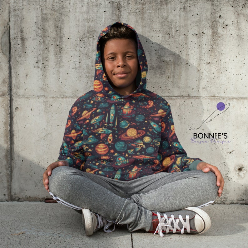 Op de afbeelding: Een jong persoon draagt een donkerblauwe hoodie met een kleurrijke print met een ruimtethema. De print bevat planeten, sterren, raketten en ruimteschepen. De hoodie heeft een capuchon met trekkoord en een kangoeroezak.