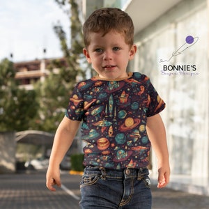 Op de afbeelding: Een jong kind draagt een blauw T-shirt met een retro print met een ruimtethema. Het shirt heeft raketten, planeten, sterren en UFO's in tinten oranje, geel, groen en blauw op een donkerblauwe achtergrond. Het kind draagt een blauwe jeans.