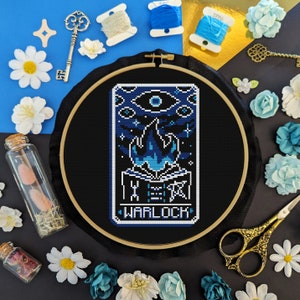 May include: A black embroidery hoop with a blue and white cross stitch design. The design features a stylized eye, flames, and a book with the word "WARLOCK" written below it.