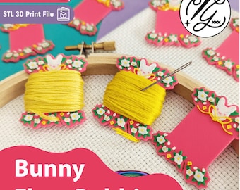 May include: Multiple pink plastic floss bobbins shaped like spools, decorated with white flowers and a white bunny head. Two bobbins are wrapped with yellow embroidery thread, one featuring a metal needle. A wooden embroidery hoop is visible.