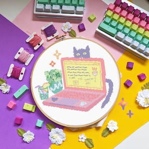 May include: A cross-stitch embroidery design featuring a black cat sitting in front of a laptop with the text "TIME TO HACK?" on the screen. The laptop is pink and purple with a white keyboard. The cat is looking at the screen. The design is surrounded by a wooden hoop.