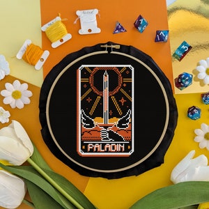 Puede incluir: Un aro de bordado negro con un diseño de punto de cruz colorido de una carta de paladín. La carta presenta una espada, un sol y la palabra "PALADIN" en naranja.