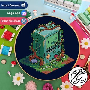 May include: A cross-stitch embroidery project in a circular wooden hoop featuring a pixelated, light blue, box-shaped character covered in green vines and small mushrooms, set against a dark navy blue background with a grid-like stitch pattern.