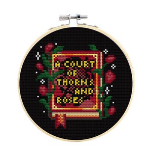 May include: A circular wooden embroidery hoop containing a cross-stitch design on black fabric. The design features a red and gold book with the text A COURT OF THORNS AND ROSES in yellow, surrounded by green vines, red rosebuds, and white stars.