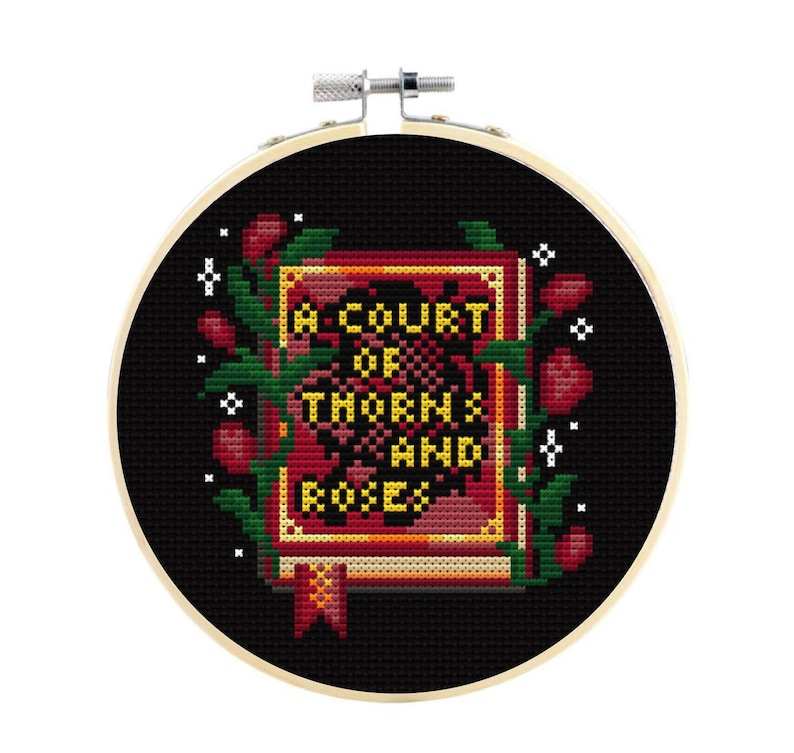 May include: A circular wooden embroidery hoop containing a cross-stitch design on black fabric. The design features a red and gold book with the text A COURT OF THORNS AND ROSES in yellow, surrounded by green vines, red rosebuds, and white stars.