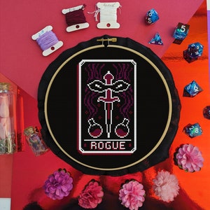 Op de afbeelding: Een zwarte borduurring met een kruissteekontwerp van een tarotkaart. De kaart is rood en zwart en heeft een zwaard en twee flesjes. De kaart staat "Rogue".