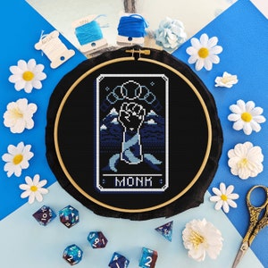 Puede incluir: Un aro de bordado negro con un diseño de punto de cruz de una mano azul sobre un fondo azul y blanco. El diseño está etiquetado como "MONK".