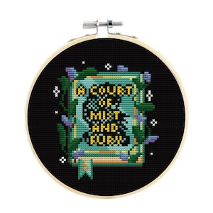 May include: A circular wooden embroidery hoop containing a cross-stitch design on black fabric. The design features a book with the text A COURT OF MIST AND FURY in yellow, surrounded by green vines, purple flowers, and small white star accents.