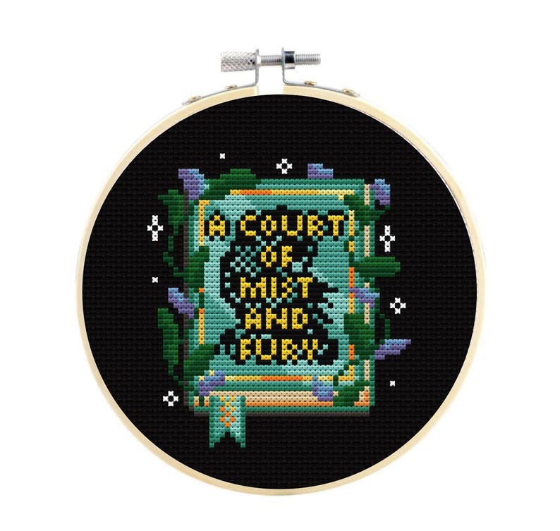 May include: A circular wooden embroidery hoop containing a cross-stitch design on black fabric. The design features a book with the text A COURT OF MIST AND FURY in yellow, surrounded by green vines, purple flowers, and small white star accents.