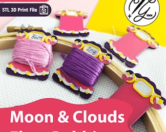May include: Several pink and purple floss bobbins shaped like clouds with yellow moon and star accents. Two bobbins are wrapped with pink and purple embroidery thread, while others are empty. The bobbins feature white labels with handwritten numbers.