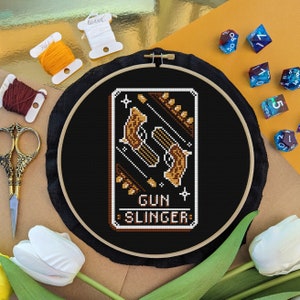 May include: A black embroidery hoop with a cross-stitch design of two gold pistols with the text "GUN SLINGER" on a black background.