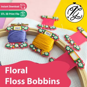Op de afbeelding: Een set roze plastic borduurgarenklosjes versierd met witte en groene bloemmotieven. Twee klosjes zijn omwikkeld met paars en geel garen, terwijl andere leeg zijn. Ze zijn gerangschikt op een witte stoffen borduurring.