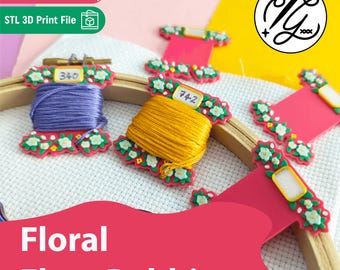 May include: A set of pink plastic embroidery floss bobbins decorated with white and green floral patterns. Two bobbins are wrapped with purple and yellow thread, while others are empty. They are arranged on a white fabric embroidery hoop.