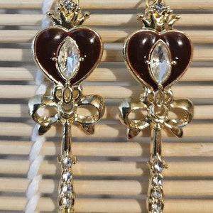 May include: A pair of gold tone earrings with a red heart design, a clear rhinestone, and a bow. The earrings are shaped like a key.