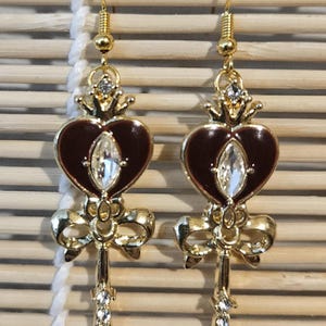 May include: A pair of gold tone earrings with a heart-shaped charm, a bow, and a key design. The heart is red with a clear rhinestone in the center. The earrings have a gold hook closure.