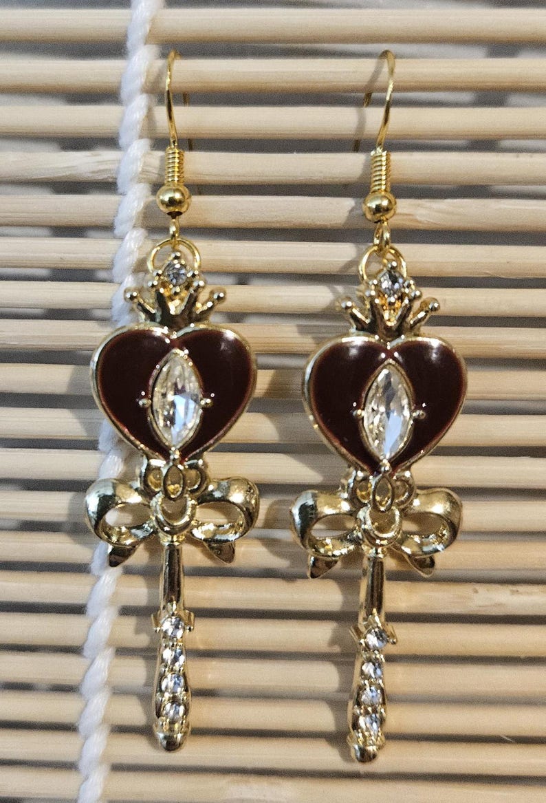 May include: A pair of gold tone earrings with a heart-shaped charm, a bow, and a key design. The heart is red with a clear rhinestone in the center. The earrings have a gold hook closure.