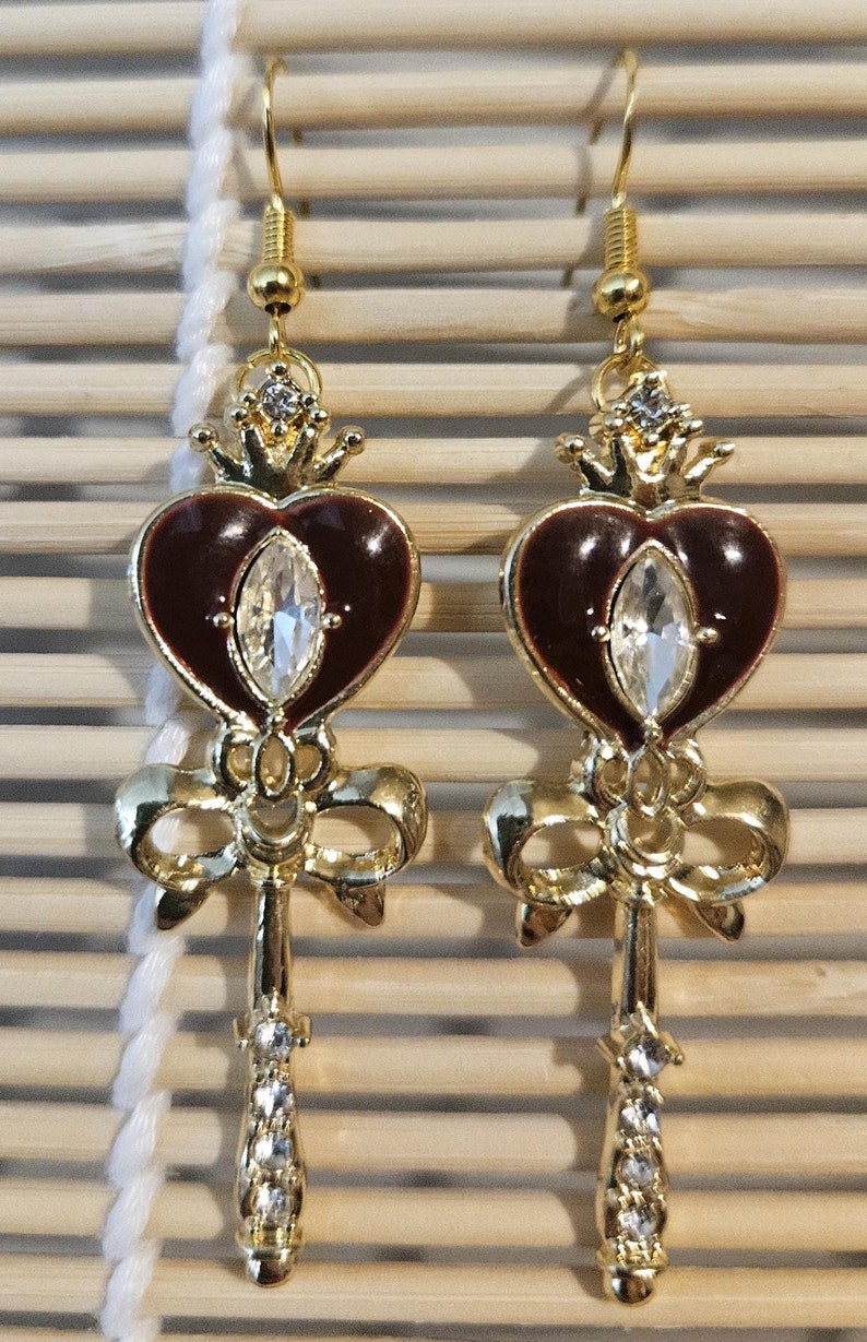 May include: A pair of gold tone earrings with a red heart design, a clear rhinestone, and a bow. The earrings are shaped like a key.