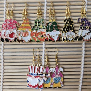 以下が含まれることがあります： 金色のフックが付いた、カラフルなノームのイヤリングのコレクション。ノームは、ハート、カボチャ、クリスマスツリーなど、さまざまなデザインが特徴です。追加のイヤリングは、愛国的なノームとウサギの耳のノームを描いています。