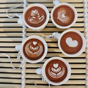 Könnte beinhalten: Fünf Paare Kaffeebecher-Ohrringe mit verschiedenen Latte-Art-Designs. Die Ohrringe sind aus weißer Keramik und haben silberne Haken.