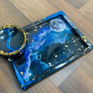 May include: A resin rolling tray and bowl set with a celestial theme. The tray and bowl are black with blue and white swirls, gold flakes, and a moon and character graphic. The bowl has a gold rim.