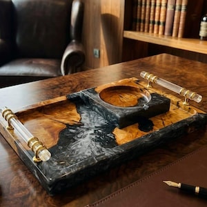 May include: A rectangular serving tray crafted from natural wood and black marbled resin, featuring two clear acrylic handles with gold-toned metal accents. A matching square-shaped ashtray with a circular center indentation sits atop the tray.
