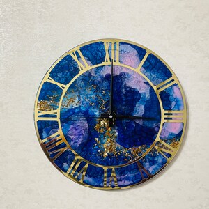 Può includere: Un orologio rotondo con numeri romani dorati. Il quadrante dell'orologio è un design astratto blu e viola con macchie dorate.