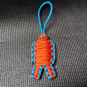 Pode incluir: Um chaveiro de paracord vermelho e azul com um laço no topo. O chaveiro é feito de cordão trançado e tem um design único.