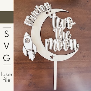 Puede incluir: Adorno de pastel de madera con las palabras "Brixton" y "two the moon" en un diseño en capas. Un cohete y estrellas también están incluidos en el diseño. El adorno está hecho de madera clara.