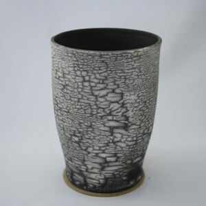 May include: A tall, cylindrical ceramic vase with a crackled glaze in shades of white and black. The vase has a wide base and a narrow opening.