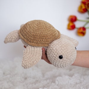 Handmade crochet turtle in a gentle brown pastel color. This turtle is big sized- 30cm! A soft and charming turtle set against a white background, with a glimpse of a few red tulips in the distance.