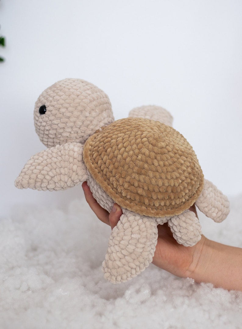 Handmade crochet turtle in a gentle brown pastel color. This turtle is big sized- 30cm! A soft and charming turtle set against a white background, with a glimpse of a few red tulips in the distance.