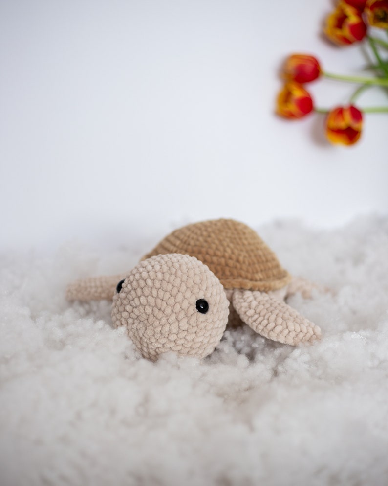 Handmade crochet turtle in a gentle brown pastel color. This turtle is big sized- 30cm! A soft and charming turtle set against a white background, with a glimpse of a few red tulips in the distance.