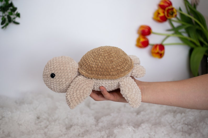 Handmade crochet turtle in a gentle brown pastel color. This turtle is big sized- 30cm! A soft and charming turtle set against a white background, with a glimpse of a few red tulips in the distance.