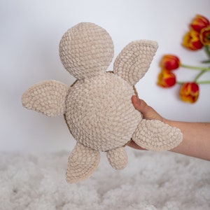 Handmade crochet turtle from the back in a gentle brown pastel color. This turtle is big sized- 30cm! A soft and charming turtle set against a white background, with a glimpse of a few red tulips in the distance.